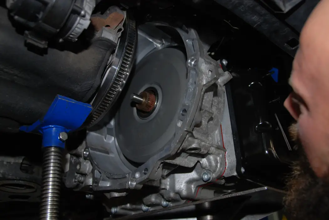 Close-up of a mechanic inspecting the torque converter and bell housing area after removing an automatic transmission from a vehicle, supported by a blue transmission jack in a workshop.