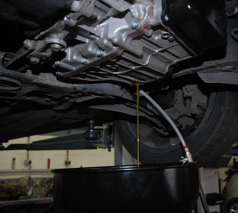 DSG transmission fluid being drained and level checked under a vehicle raised on a lift, showing the gearbox with fluid flowing through a hose into a black oil collection pan in a professional workshop.